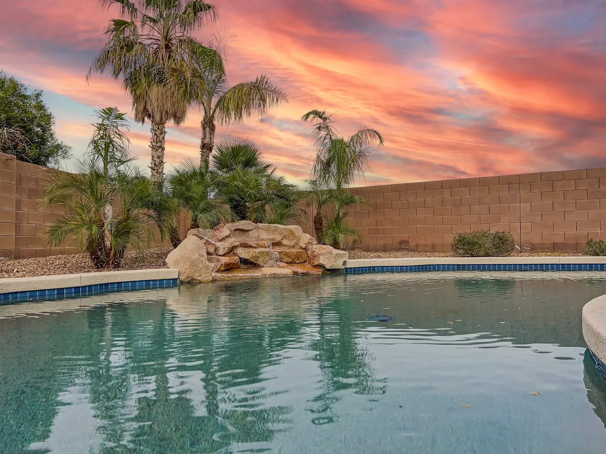 A beautiful sunset scene by the swimming pool with a waterfall feature, lush palm trees, and a relaxing patio area. The sky is glowing with sunset colors, creating a serene atmosphere perfect for evening relaxation or social gatherings.
