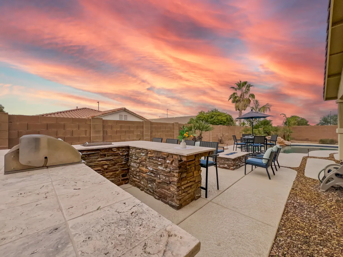 A view of an outdoor kitchen and dining area adjacent to the swimming pool, all bathed in the warm hues of a sunset sky. The space is set up for barbecuing and alfresco dining, with comfortable seating that encourages social gatherings and meals under the stars.
