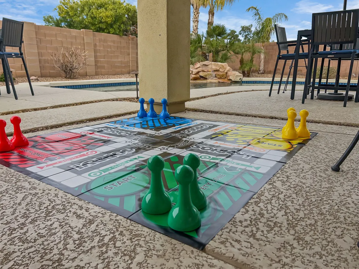 This image features a lively outdoor pool area surrounded by lush greenery. In the foreground, a board game with colored pawns sits on a table, suggesting playful, communal outdoor activities. The setting is vibrant and welcoming, with ample space for relaxation and fun in a secure, well-maintained environment.