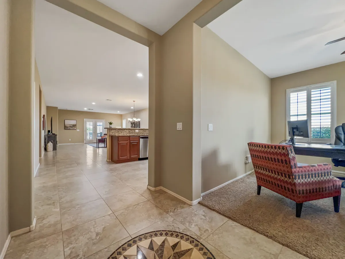 This image reveals a welcoming, multi-purpose interior where the home office area transitions seamlessly from the dining and kitchen zones. Natural light pours in, and a desk and decorative chair are positioned by the window, highlighting a pleasant workspace. The kitchen and dining area in the background subtly reinforce that the home office is set apart, yet close to daily living amenities.