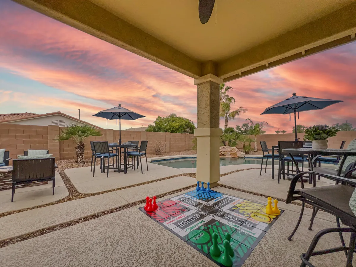 This image showcases an inviting outdoor seating area beside the pool, set against a tranquil sunset sky. The scene includes a game board, indicating space for friendly competition and group fun, while the spacious layout encourages gatherings and relaxation. The pool and surrounding greenery evoke a sense of vacation and leisure.