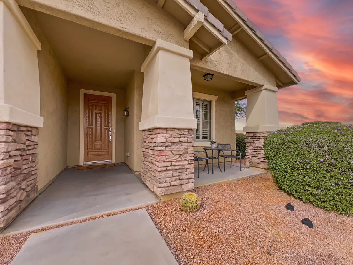 The front entrance of the home features stone columns, a welcoming front door, a small table and chairs, and manicured bushes. The peaceful seating area suggests a perfect spot for morning coffee or winding down at sunset.