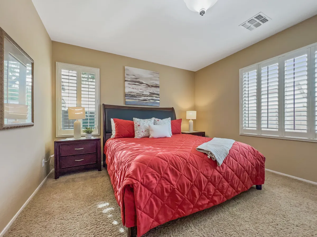 A cozy bedroom with a red bedspread, decorative pillows, and artwork on the wall. A nightstand with a lamp offers warm lighting, creating an inviting atmosphere ideal for resting and reading.