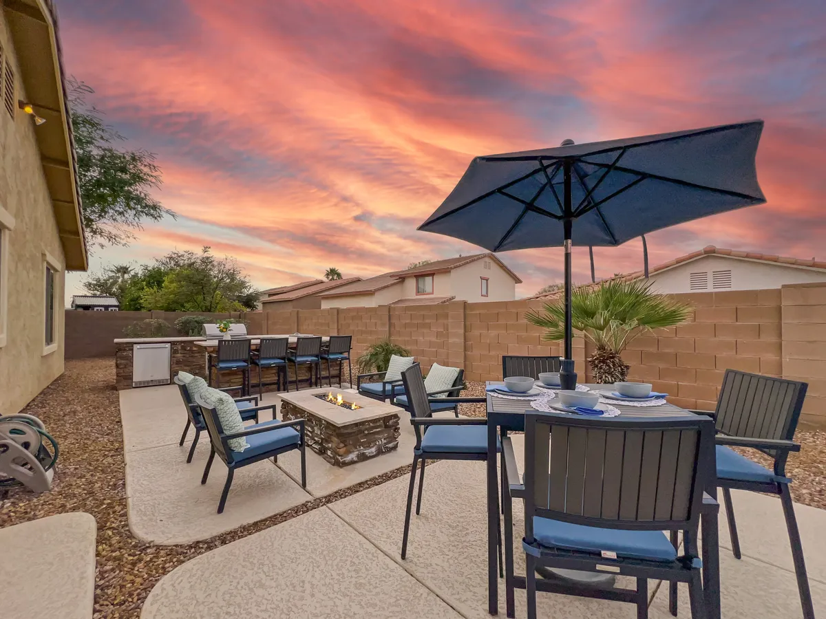 An expansive outdoor dining table sits near a cozy fire pit under an umbrella, with the sky painted in warm sunset hues. The setup is ideal for al fresco dining, celebrations, and unwinding with friends as day turns to night.