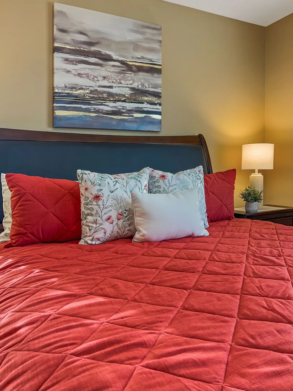 A tranquil bedroom featuring a red quilted bedspread, decorative pillows, abstract wall art, and a bedside lamp with a small plant. The overall mood is peaceful and perfect for relaxation or quiet time.